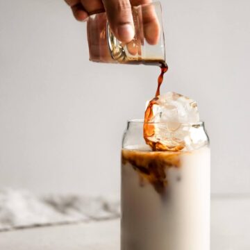 Hand pouring espresso shot into a glass of iced almond milk to make an iced vanilla latte..