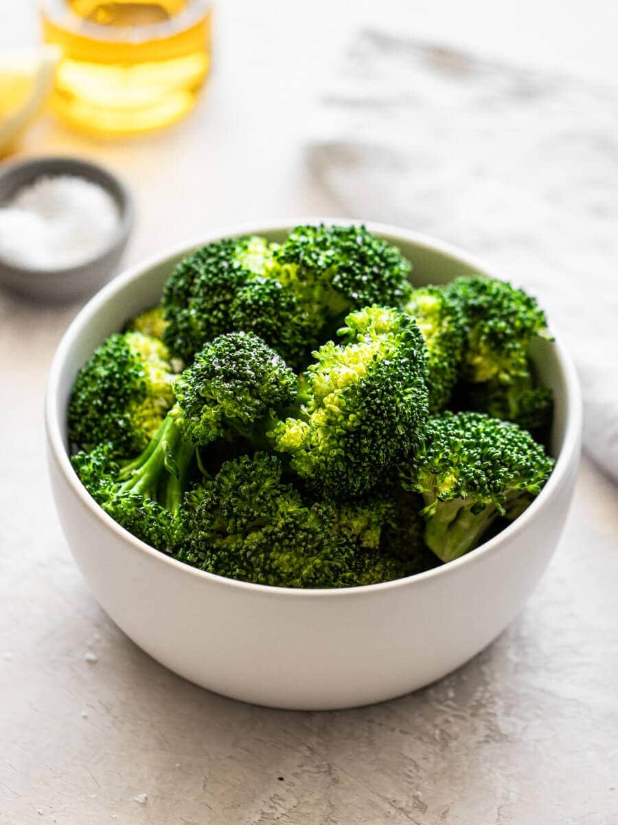 A bowl of cooked broccoli with a small bowl of flaky salt in the background.