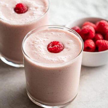 Two glasses of smoothie with a bowl of raspberries.