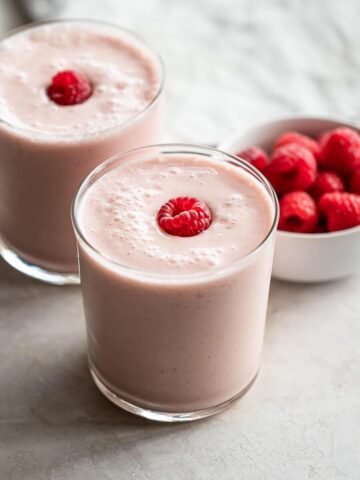Two glasses of smoothie with a bowl of raspberries.