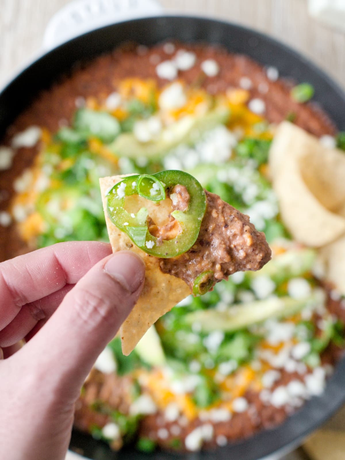 Hand holding a tortilla chip with bean dip and slice of jalapeno.