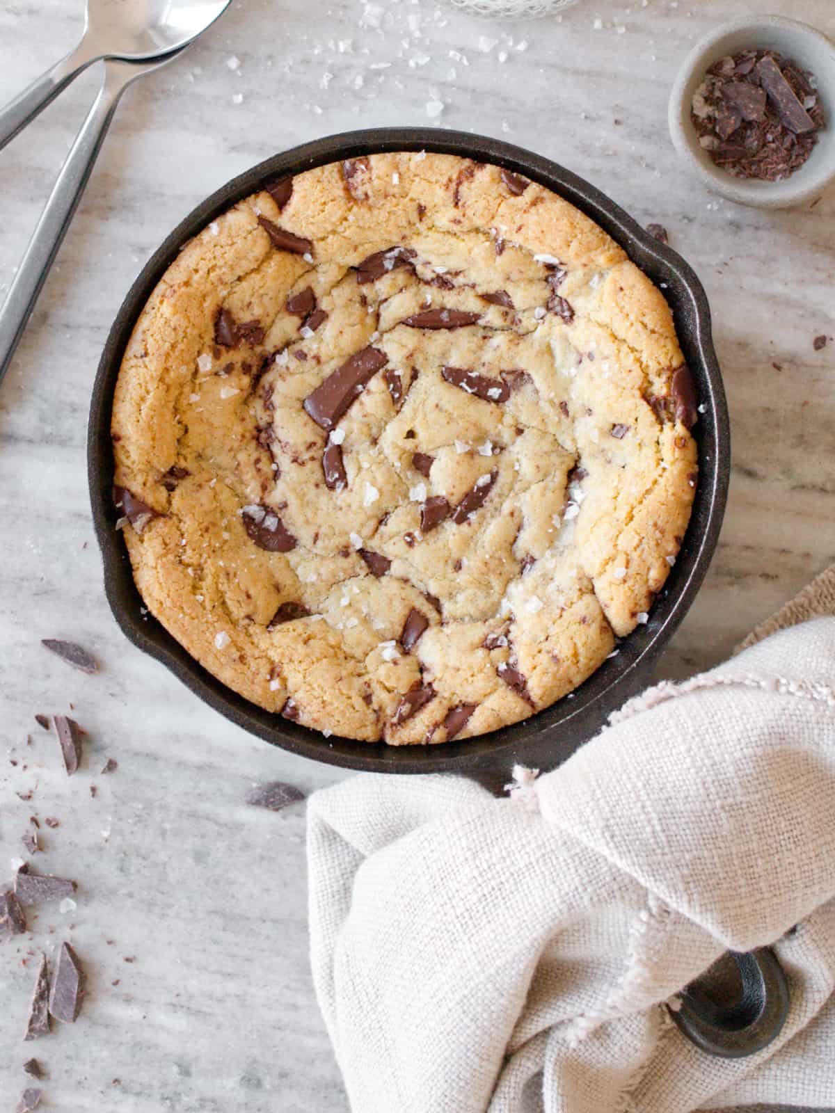 Baked mini skillet cookie with towel, bowl of chocolate chunks, and two spoons.