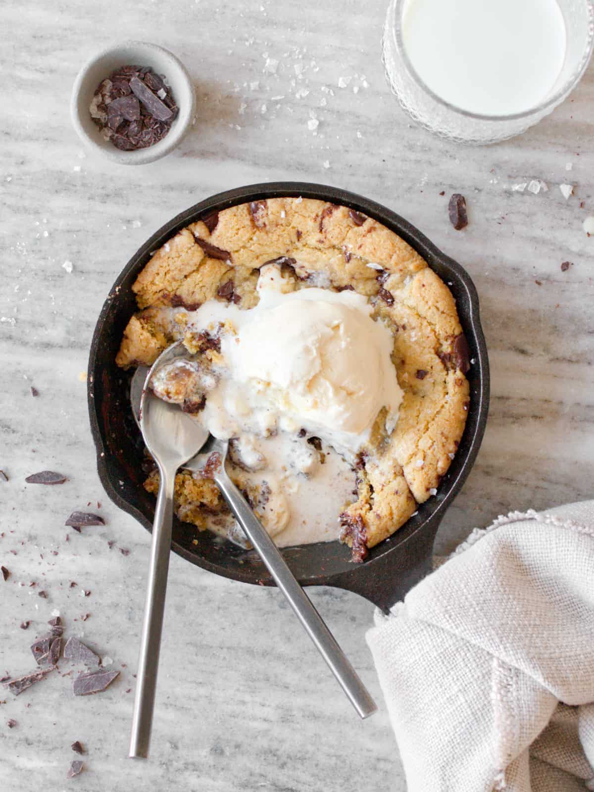 Half eaten mini skillet cookie with melting ice cream, two spoons, and a glass of milk.