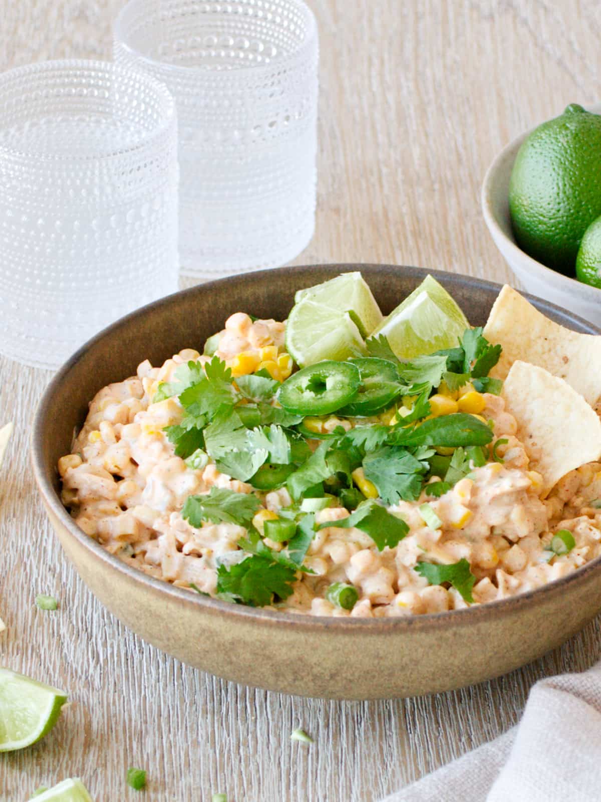 Bowl of corn dip with two glasses of drinks and a bowl of limes.