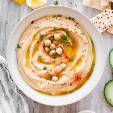 Bowl of hummus surrounded by cucumber, towel, pita bread, and lemon.
