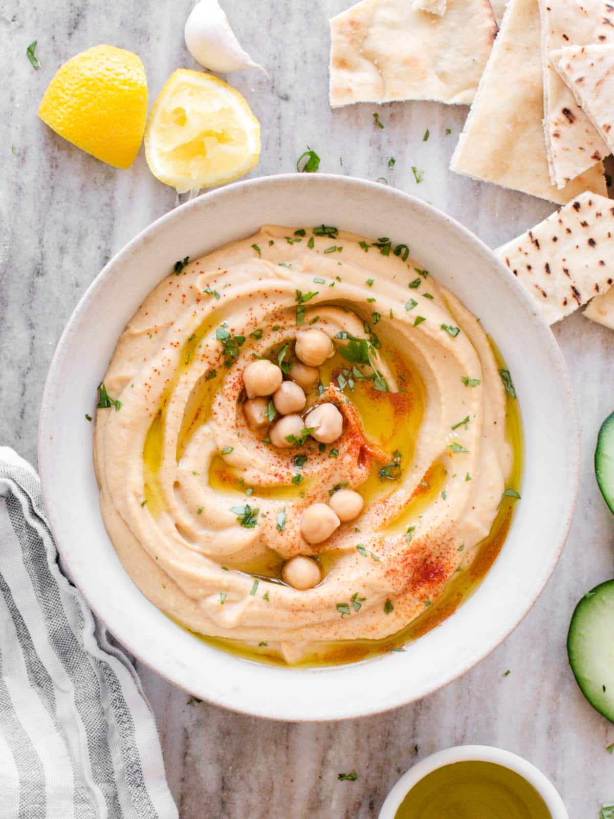Bowl of hummus surrounded by cucumber, towel, pita bread, and lemon.